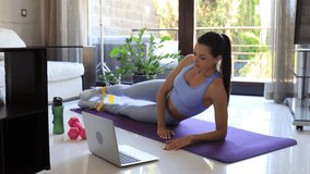 Fitness beautiful slim woman doing side plank and watching online tutorials on laptop, training in living room. Stay at home activities.