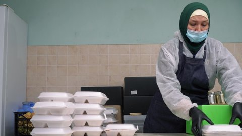 Eid al-Adha or the Feast of Sacrifice. Islamic relief and Aid. Muslim woman volunteer wearing a hijab, mask and gloves. Cooking food, packaging of hot meals in lunch boxes, distribution to the needy