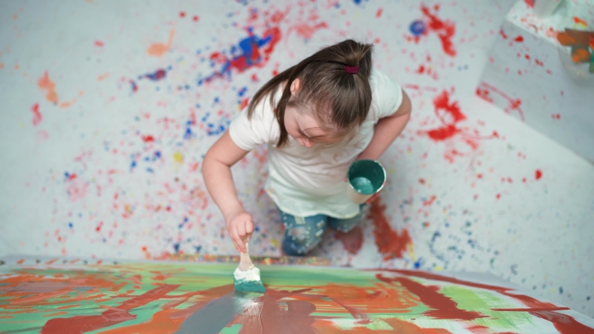 Girl with down syndrome draws with a brush on a large canvas in a white room, girl with special needs draws a color abstraction, top view, 4k slow motion. Royalty-Free Stock Footage #1075025549
