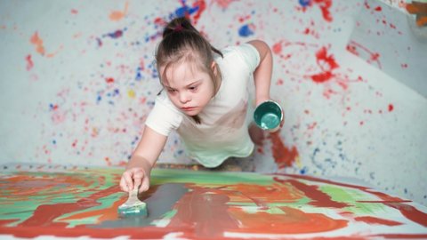 Girl with down syndrome draws with a brush on a large canvas in a white room, girl with special needs draws a color abstraction, top view, 4k slow motion.