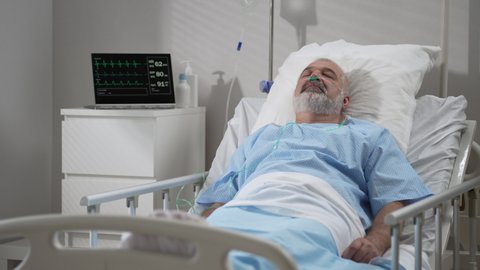 An elderly patient wakes up coming out of a coma. Open your eyes while lying on a bed in a hospital connected In the Hospital Sick Male Patient Sleeps on the Bed, .