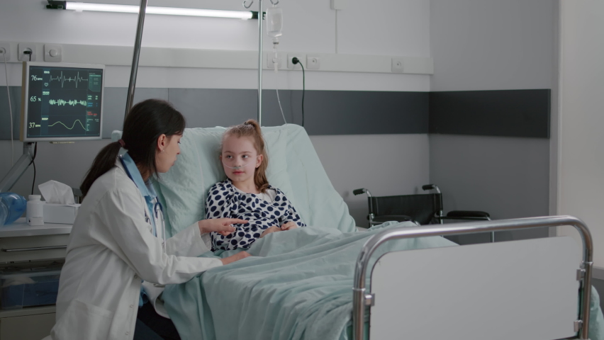 Pediatric woman doctor interacting with sick patient giving high five during clinical consultation in hospital ward. Child lying in bed wearing oxygen nasal tube waiting for medication treatment | Shutterstock HD Video #1075036892