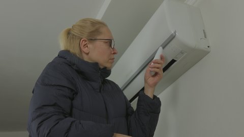 A woman dressed in a warm jacket tries unsuccessfully to turn on the air conditioner in the apartment.