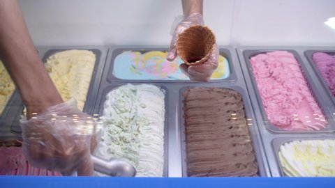 Close-up of a showcase with different flavors of Italian ice cream. The seller takes a delicious cold dessert with a spoon and puts the ice cream in a waffle cup.