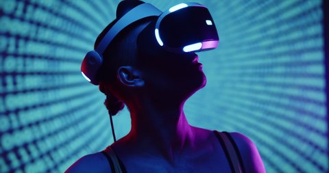 Cyber gamer in VR glasses plays virtual reality game in neon futuristic space. Cyberpunk fashion gaming concept. Young handsome man looks around and shooting from virtual blaster gun.