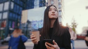 Young attractive chinese business girl walking in city center. Cheerful carefree woman drinking takeaway coffee and texting mobile phone messages. - Powered by Shutterstock - Get 15% off with code: PIKWIZARD15