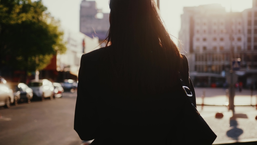 Following young tourist beautiful woman wearing black business jacket and bag, enjoying sunlight in big city, walking down the street.