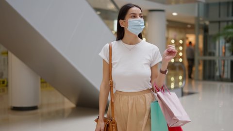 Young brunette woman in medical mask with different shopping paper bags looking on glass showcases with clothes while walking in mall, feeling pleased and glad to spend money after covid19 quarantine