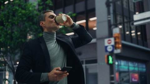 Energetic Handsome Young Adult Businessman Walking Fast, Drinking Take Away Coffee and Using Smartphone on the Street in a City. Confident Executive Connecting with People Online His Way to Office.