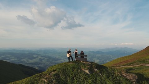 Aerial view of three people enjoying on their mountains vacation at sunset. Musician playing guitar and piano in the mountains. Singer vocalist woman, pianist, guitarist man playing song performing.