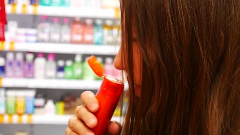 A cute long haired girl chooses shampoo opening the red tube and sniffing it in the store close-up
