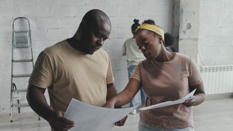 Medium shot of African-American man and woman looking at home remodeling plans and chatting while kids measuring wall in background