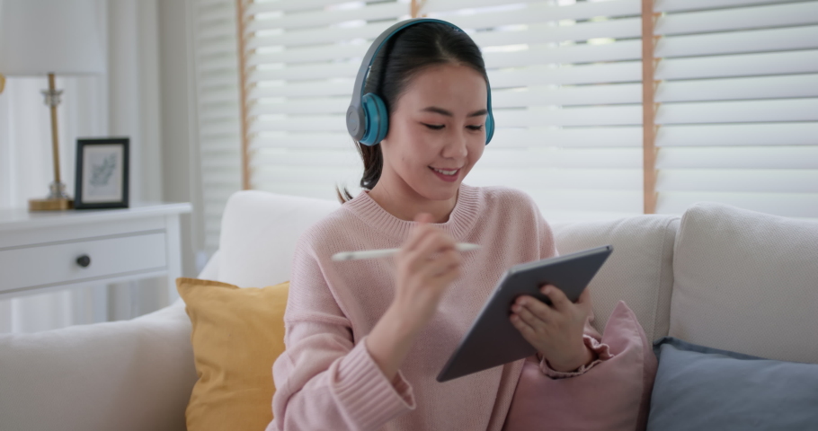 Young teen asia happy people lady girl watch live event web video call wear wireless headset write idea diary on digital wifi note pad app sit on sofa couch at home in reskill upskill mba exam class. | Shutterstock HD Video #1075048595