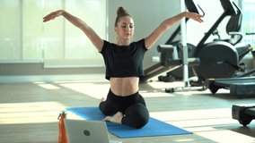 young fit woman in sportswear doing  yoga on mat watching  video online training on laptop computer in fitness gym . sporty female practicing online sport class stretching and exercises . workout 
