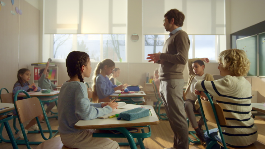 Mixed race group of pupils learning in school classroom. Male teacher asking questions at lesson. Crane shot of active students raising hands. Young schoolteacher talking with children in class Royalty-Free Stock Footage #1075050548