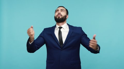 Positive cheerful young bearded man in official style suit having fun dancing to music and singing song, resting and enjoying on party. Indoor studio shot isolated on blue background.
