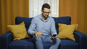 Smiling man in shirt with smartphone making video call while sitting on couch in living room. Portrait of happy young male mobile video calling, sitting on sofa at home