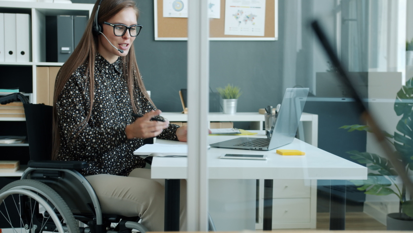 Cheerful handicapped girl employee is talking to clients online with laptop and headphones laughing sitting in wheelchair in modern office. Business and disability concept. | Shutterstock HD Video #1075060340