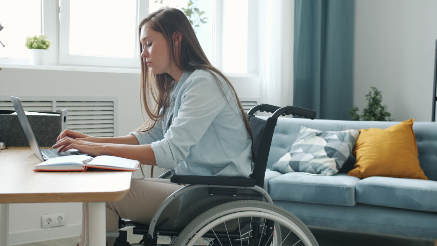 Motivated handicapped freelancer is working with laptop typing and taking notes sitting in wheelchair at home. Distant work and disability concept. Royalty-Free Stock Footage #1075060418
