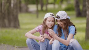 Children friends use smartphone, play games or watch videos on mobile phone screen. Girlfriend sisters are sitting nearby in park and watching something on a smartphone
