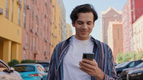 Positive Young Eastern Man in stylish shirt walking and enjoys Chatting on Phone. Smiling Mixed Race Guy Scrolling Posts or Watching Video using Smartphone Applications. Media. Colorful Buildings.