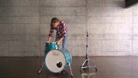 WIDE shot of Caucasian bearded long haired man is Setting Up A Drum Set  before Playing - Drum Lesson. 