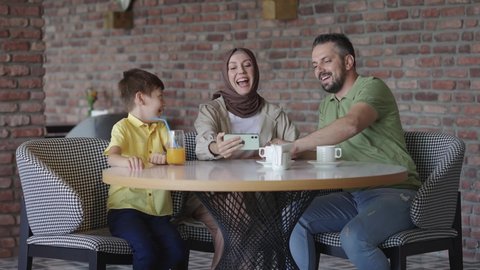 Stylish hijab mother, bearded father and their children are watching a funny video with smartphones in cafe. Watching videos on the phone. The concept of having fun as a family.