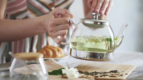 Уoung girl puts glass teapot on bamboo board with dry green tea foxes and sprig of jasmine. Fresh jasmine flowers drenched in hot water. Making flower tea on sunny morning. Croissant in background.
