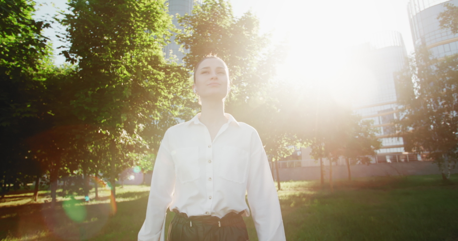 Happy business woman walking in green garden by eco friendly office building backlit with bright sunlight. Smiling independent female enjoying successful corporate career, wide angle following shot