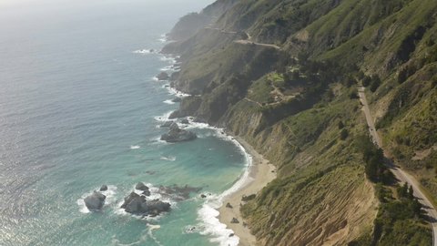 Aerial parallax view of rocky Big Sur beach and highway in green scenic hills