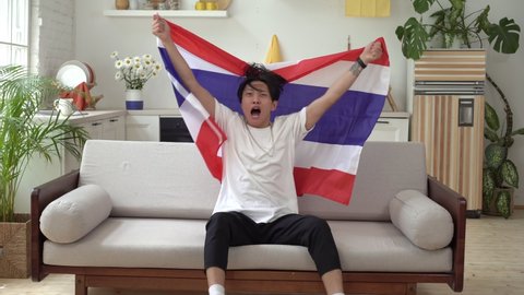 Thai fan watching sports at home on TV. An Asian man with the flag of Thailand supports the national team