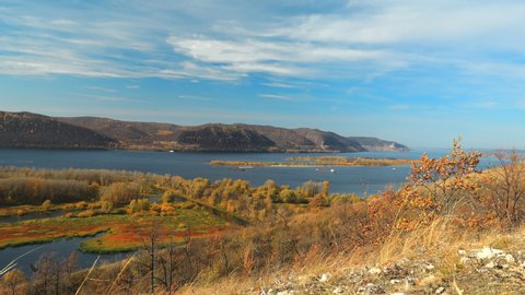 Wide river flowing between high hills filmed in time lapse. The Volga river near the Zhigulevsky mountains. Autumn landscape. Orange colors of nature under the blue sky. Steamers move along the river.
