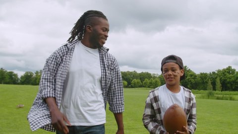 Happy handsome african american father with dreadlocks and cute preadolescent son with eye black holding american football ball, talking and walking through green filed to play football outdoors.