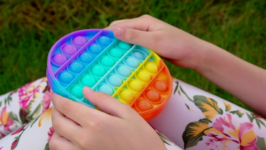 Child is playing trendy popular game anti-stress toy pop it. Little girl is sitting on grass and playing popit.