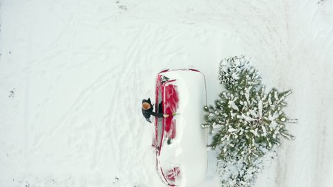 Woman cleans the roof of a car with a brush from snow on a sunny winter day. Top-down view of a snow-covered car and a young woman clearing the snow from the car.