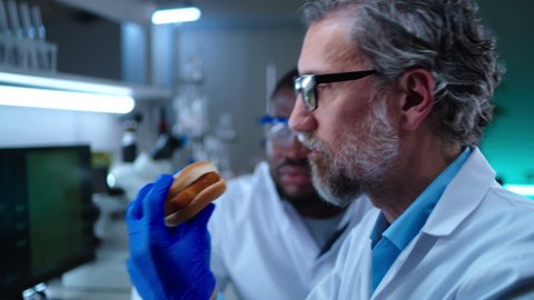 Rack focus of mature male scientist biting and chewing burger with cell lab grown meat and discussing taste with African American coworker in laboratory