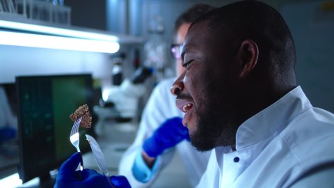 Rack focus of African American man microbiologist eating piece of patty made of cell cultured lab grown meat and speaking with mature coworker in lab