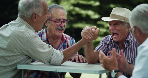 Cinematic shot of cheerful senior retired friends are having fun to play arm wrestling and laughing together in green park. Concept of retirement, active lifestyle, elderly, friendship, competition