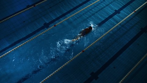 Female Swimmer Racing in Swimming Pool. Professional Athlete Determined to Win Championship using Butterfly Style. Dark Dramatic Colors, Cinematic Lap Lane Light. Aerial Tracking Dutch Angle Shot
