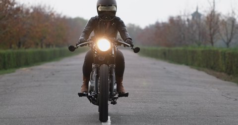 Woman biker in black helmet and leather jacket rides motorcycle with head-light along asphalt road approaching camera in evening