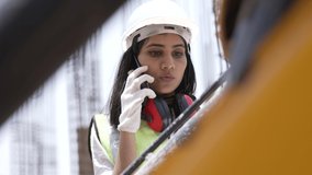 Young attractive female Indian civil engineer in safety jacket and helmet is talking on a mobile phone standing of under development building near the construction site outdoor.Asian engineer on site. - Powered by Shutterstock - Get 15% off with code: PIKWIZARD15