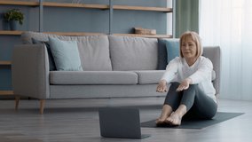 Online Fitness. Senior Lady Doing Seated Straddle Exercise At Laptop Computer Stretching Trying To Touch Toes Sitting On Floor At Home. Older Lady Exercising Having Distant Workout Near Computer