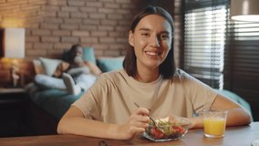 Young beautiful woman eating vegetable salad and chatting at camera via online video call while staying at home with husband during isolation