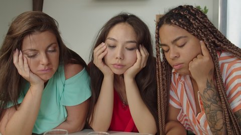 Close-up of glasses and portraits of three multiracial women in colorful clothing suffering from hangover, dropping painkiller pill in water to relieve headache painful feelings at domestic kitchen.