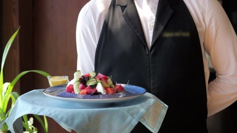 The waiter carries a plate with a delicious salad along the panoramic windows to the client. A waiter in a white shirt and a black apron, serving on the go, in a modern restaurant. Customer service