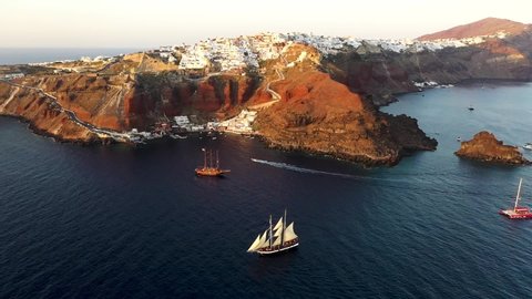 Aerial drone view video of beautiful sail boats cruising in the deep dark blue Aegean sea during sunset, Santorini, Cyclades, Greec