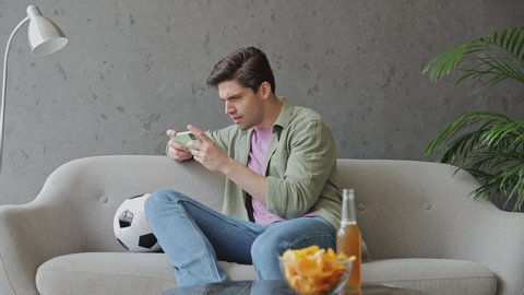 Young sad upset man fan in green clothes t-shirt cheer up support sport football team sit on grey sofa with soccer ball bottle beer eat chips wait watch tv on mobile cell phone indoors room gray wall