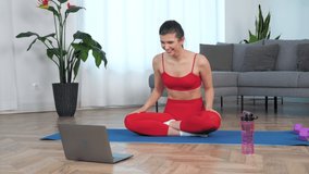 Sportive smiling woman in sportswear sitting on yoga mat makes wave with hands at home. Positive fitness girl doing exercise training workout, study watch online sport webinar lesson laptop computer