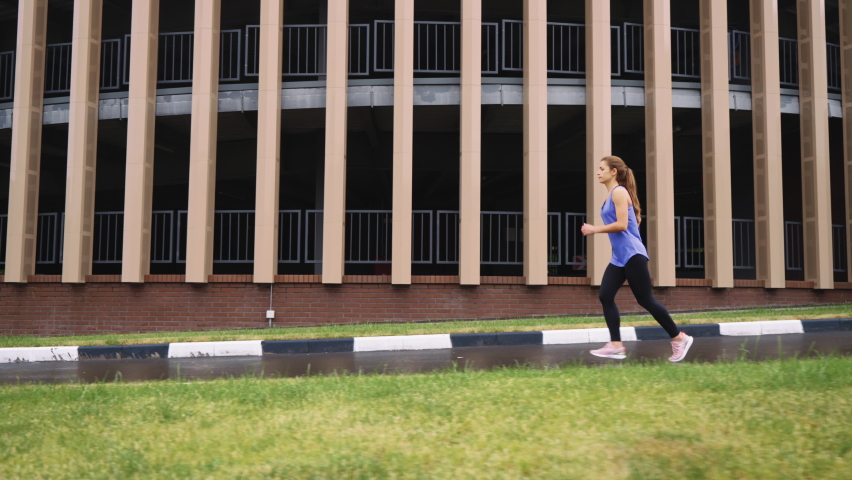 Slow motion woman in sportswear jogging against rounded building, wet ground after rain. Tracking shot female athlete training in city early in the morning. Concept of fitness