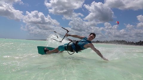 SLOW MOTION, CLOSE UP: Happy young man kiteboarding in Tanzania drags his hand along the surface of the crystal clear ocean. Fit male kiteboarder surfs around the blue lagoon in stunning Zanzibar.